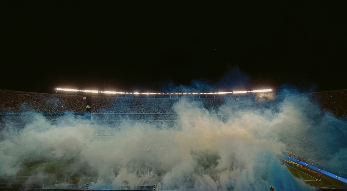 Screenshot 2026-08-06 alle 19.27.05 Vista panoramica di uno stadio di calcio illuminato di notte, parzialmente coperto da una fitta nube di fumo azzurro e bianco creata dai tifosi sulle tribune.